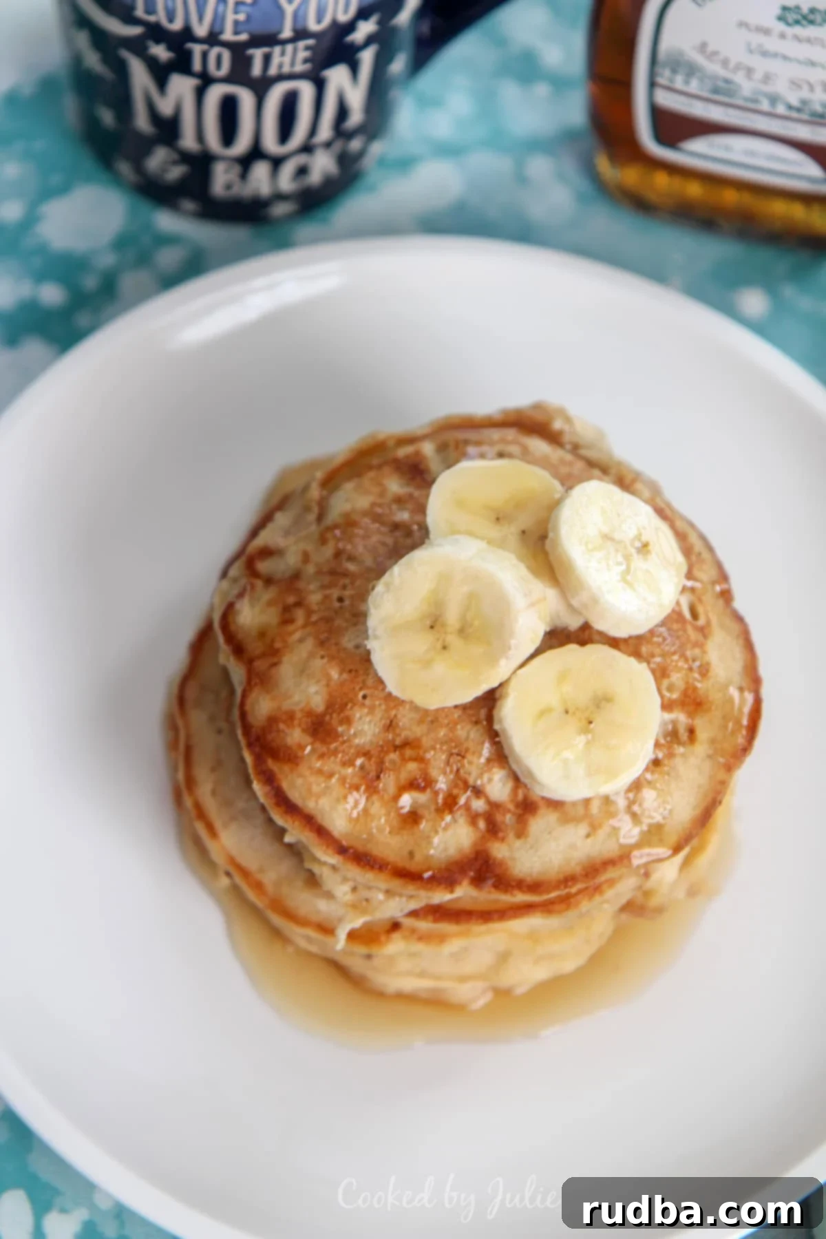 A tall stack of golden banana pancakes on a white plate, adorned with sliced fresh bananas and drizzled with glistening maple syrup.