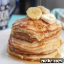 Six fluffy banana pancakes on a white plate with fresh banana slices on top, a generous drizzle of maple syrup, and a blue mug in the background.