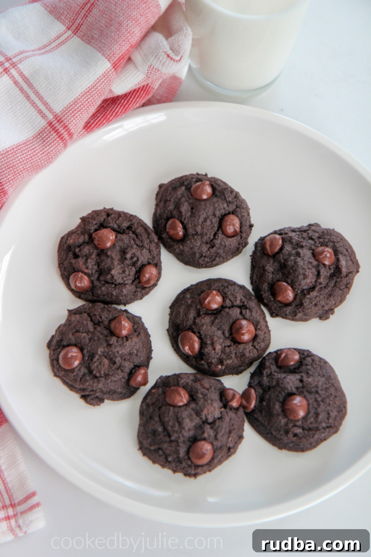 A stunning arrangement of freshly baked double chocolate cookies on a white plate, accompanied by a tall glass of cold milk and a charming red and white checkered towel.
