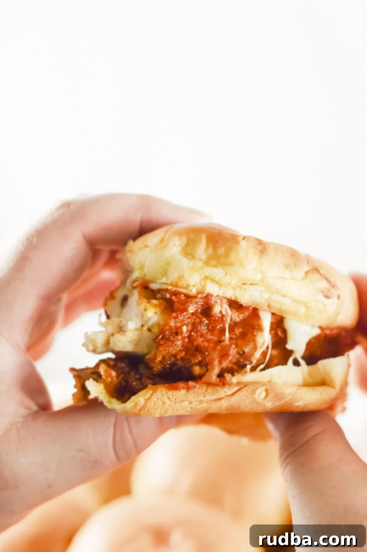 Two hands holding up a single chicken parm slider, perfectly highlighting the crispy golden chicken cutlet, the rich melted cheese, and the soft, lightly toasted bun, inviting viewers to take a bite.