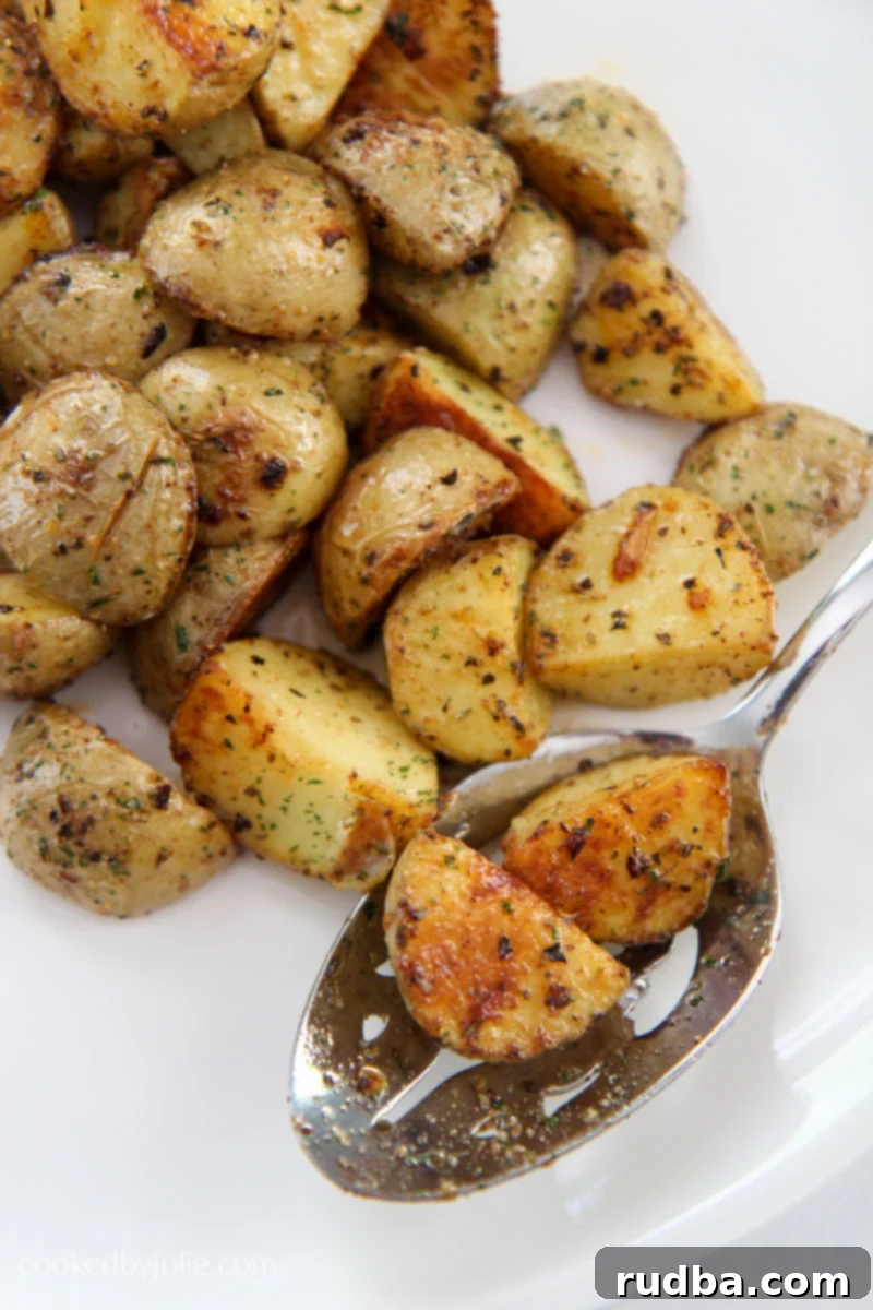 Two perfectly roasted garlic potatoes resting on a slotted spoon, with a pile of more golden, crispy potatoes surrounding it on a white plate, highlighting their appetizing texture.