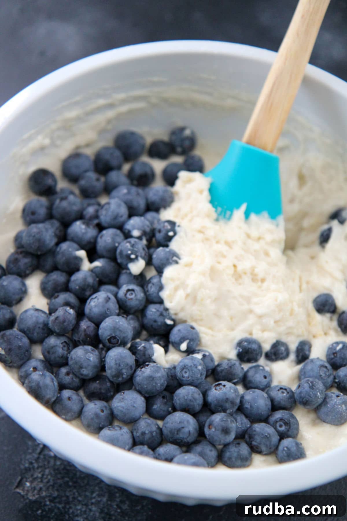 Bowl of thick lemon blueberry pancake batter with a spatula, with fresh blueberries and lemon slices arranged beside it on a rustic surface.