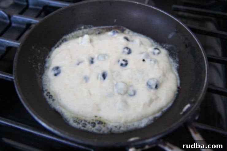 A golden brown lemon blueberry pancake cooking in a black cast-iron skillet, with visible blueberries embedded in the batter.