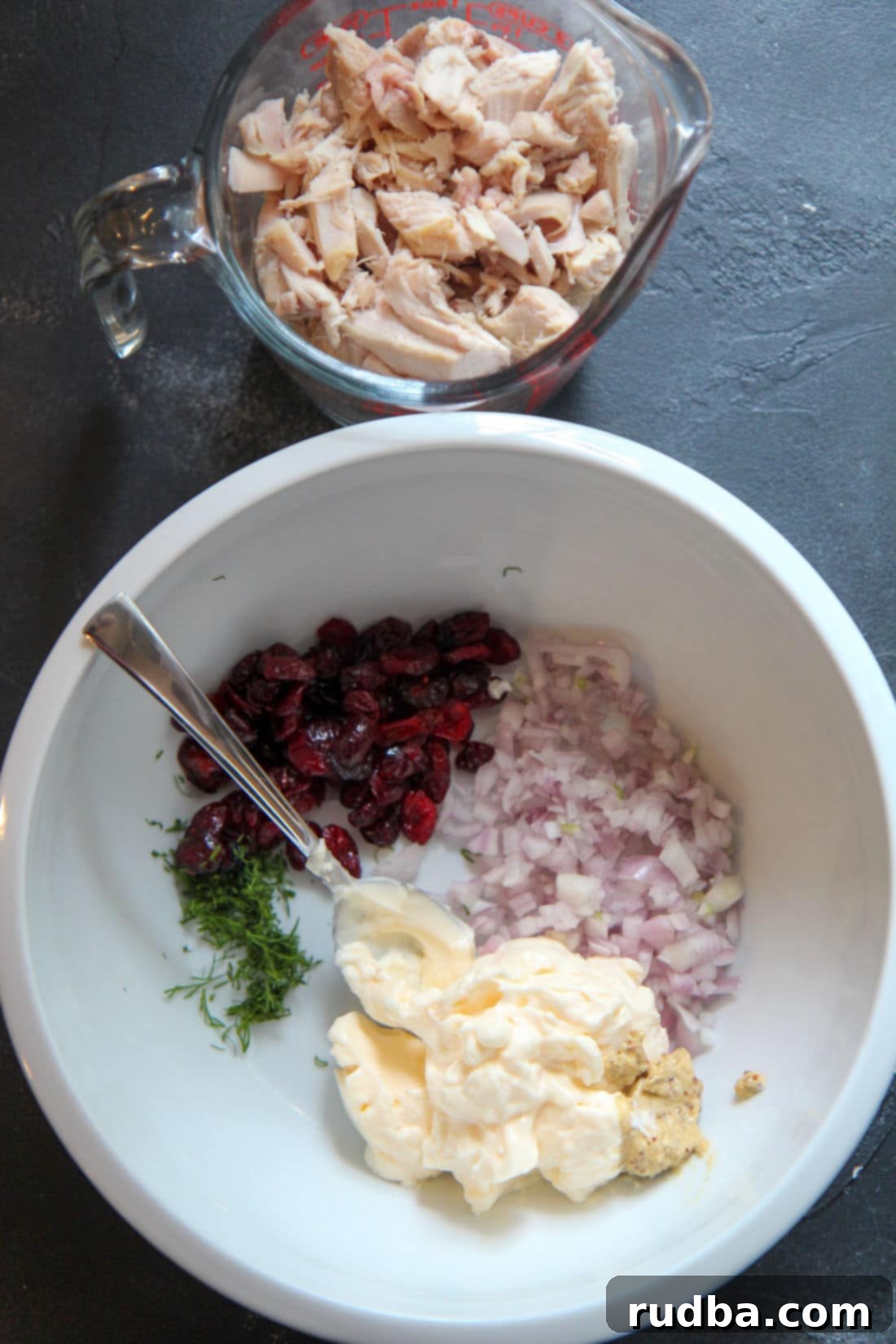 A vibrant close-up of individual chicken salad ingredients laid out in small white bowls: creamy mayonnaise, finely diced shallots, plump dried cranberries, and fresh green dill, with diced cooked chicken in a measuring cup, all poised for mixing.