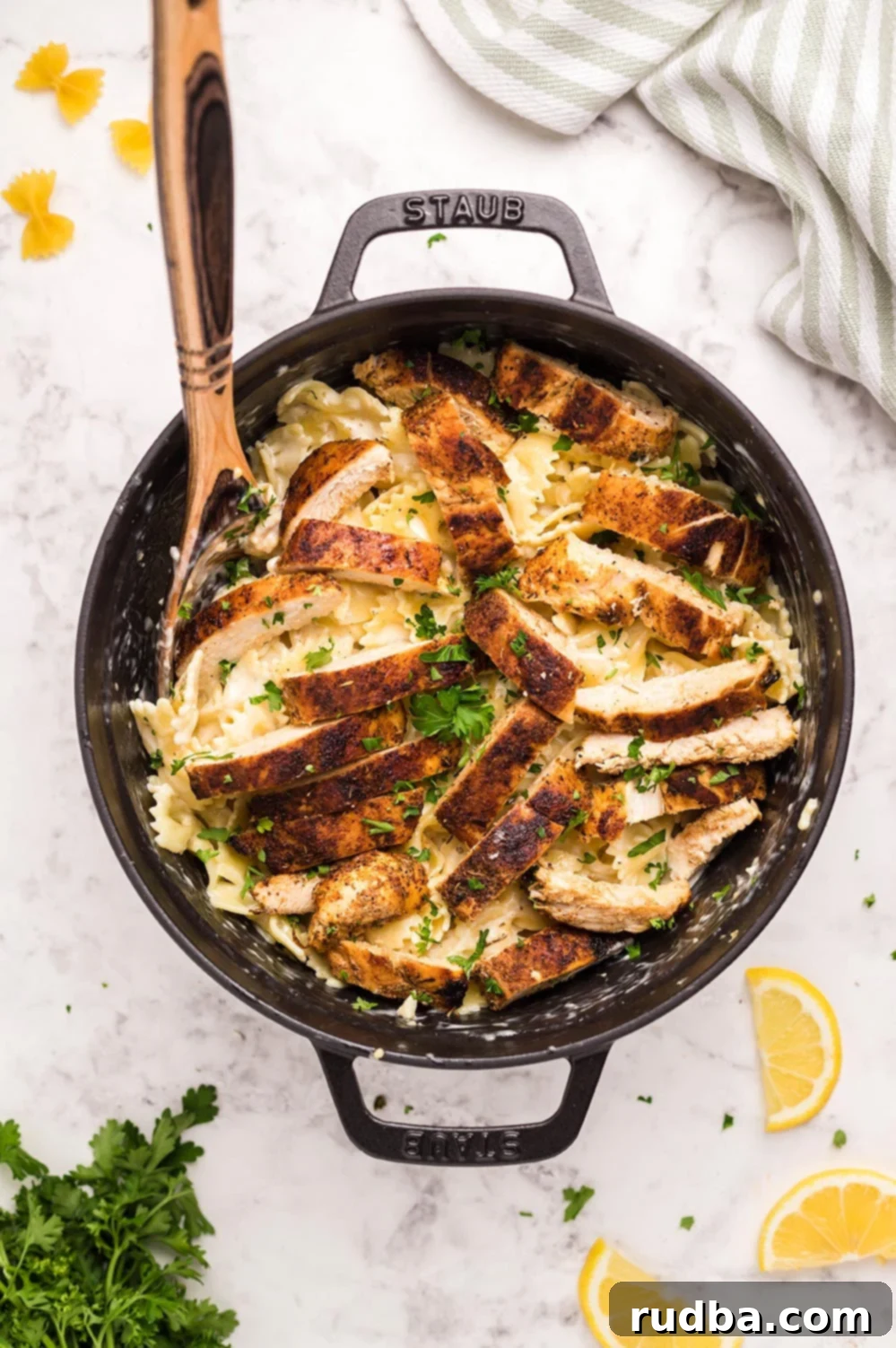 Cajun chicken, farfalle pasta, parsley, and a wooden spoon in a black skillet, showcasing a mouthwatering dish ready to be served.