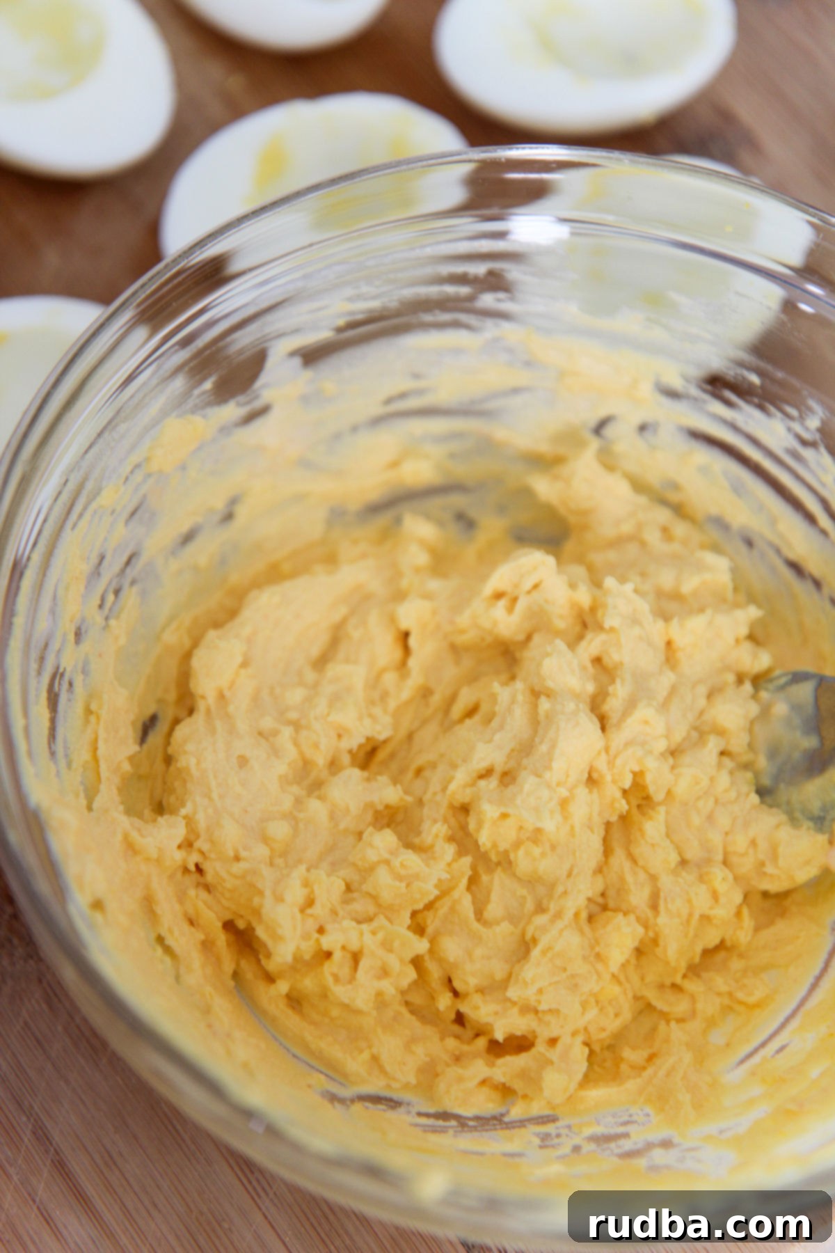 Close-up of creamy egg yolk mixture in a clear bowl with hard-boiled egg whites blurred in the background, ready for assembly.