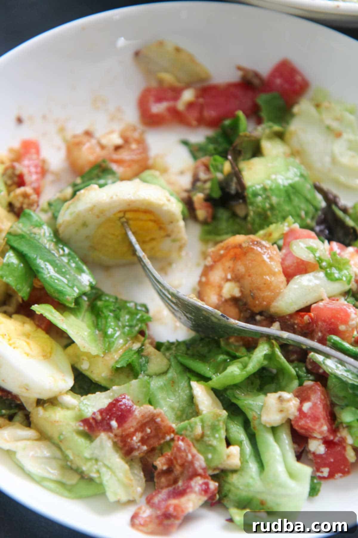 A fork holding a piece of hard-boiled egg over a plate of colorful shrimp cobb salad.