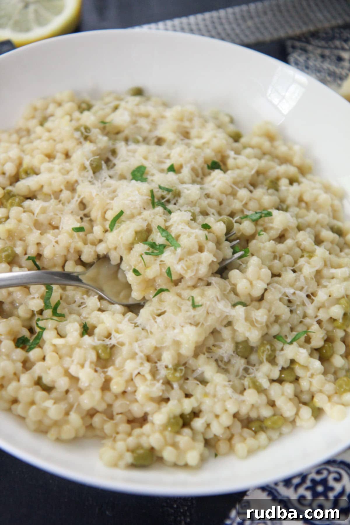Close-up of fluffy lemon pearl couscous in a white plate with a fork, garnished with fresh parsley.