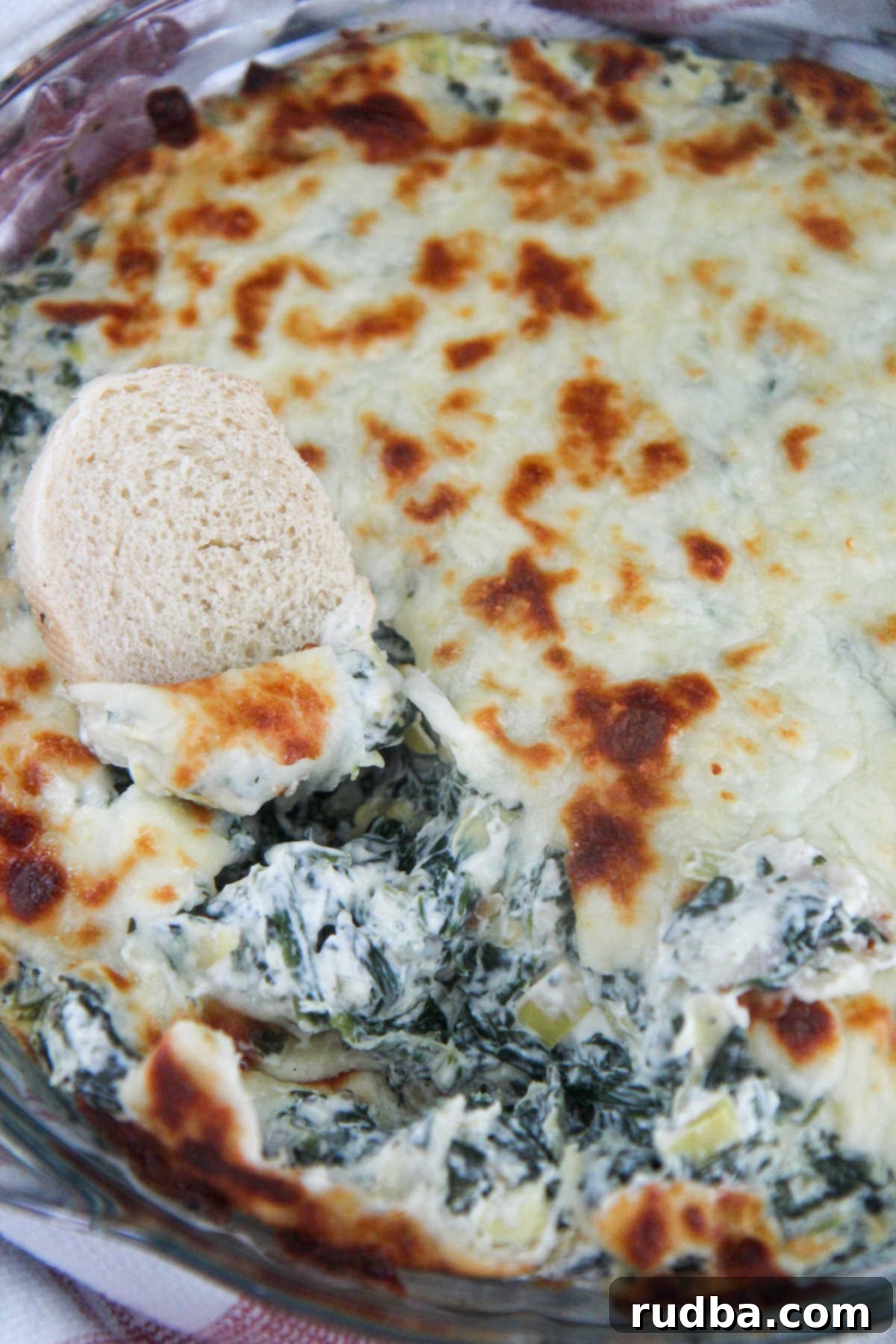 A close-up of creamy spinach artichoke dip in a clear glass bowl, with pieces of bread ready for dipping.
