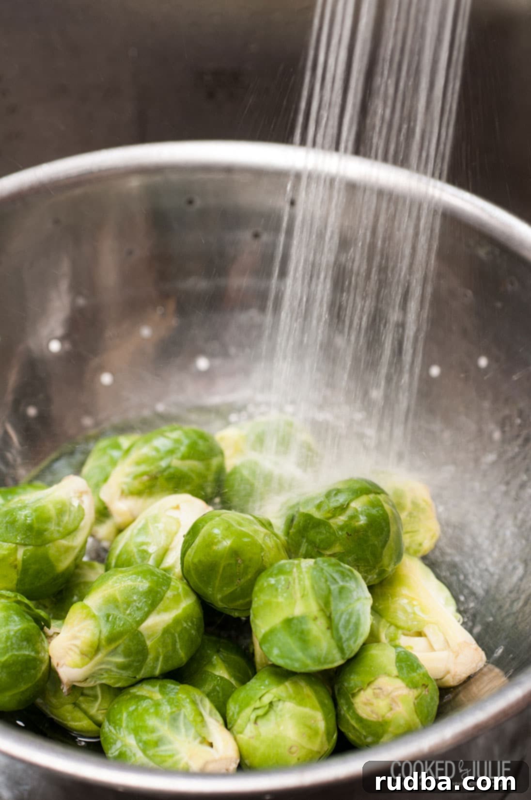 Fresh Brussels sprouts being washed under cold running water in a metal colander