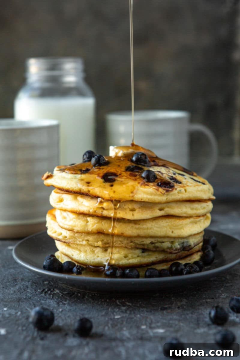 six blueberry pancakes on a blue plate with butter and maple syrup on top. A jug of milk in the background with two mugs and fresh blueberries.