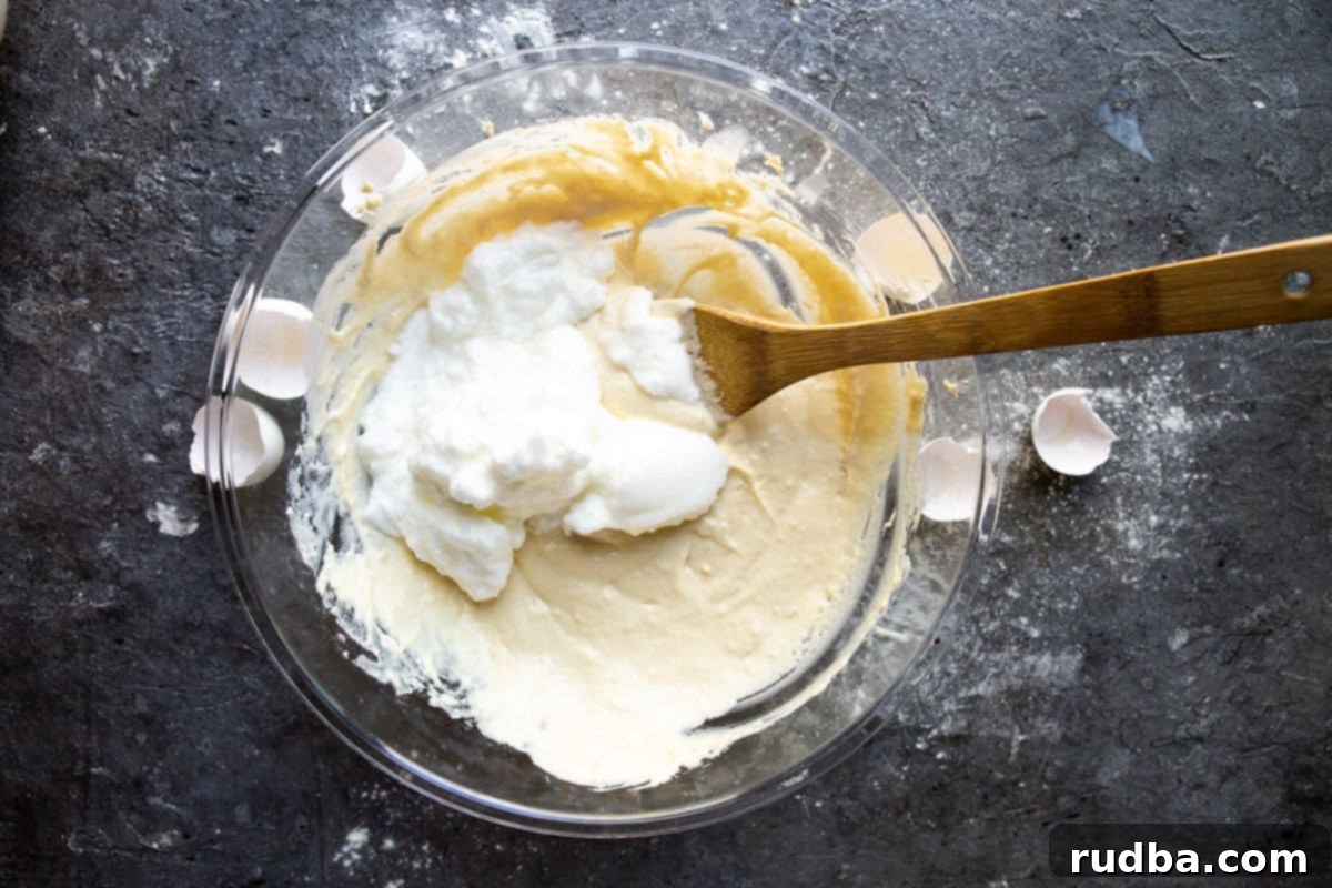 A bowl with pancake batter and folded egg whites with a spatula. Egg shells on the side of the bowl. 