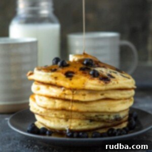 Fluffy Blueberry Pancakes 7 six blueberry pancakes on a blue plate with butter and maple syrup on top. A jug of milk in the background with two mugs and fresh blueberries.