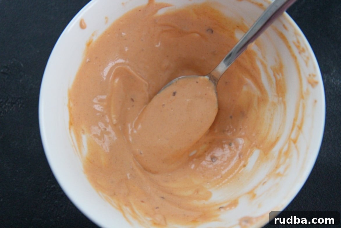 A close-up of a white bowl overflowing with a rich, orange-hued chipotle mayo sauce, with a spoon resting inside, ready for dipping or spreading.