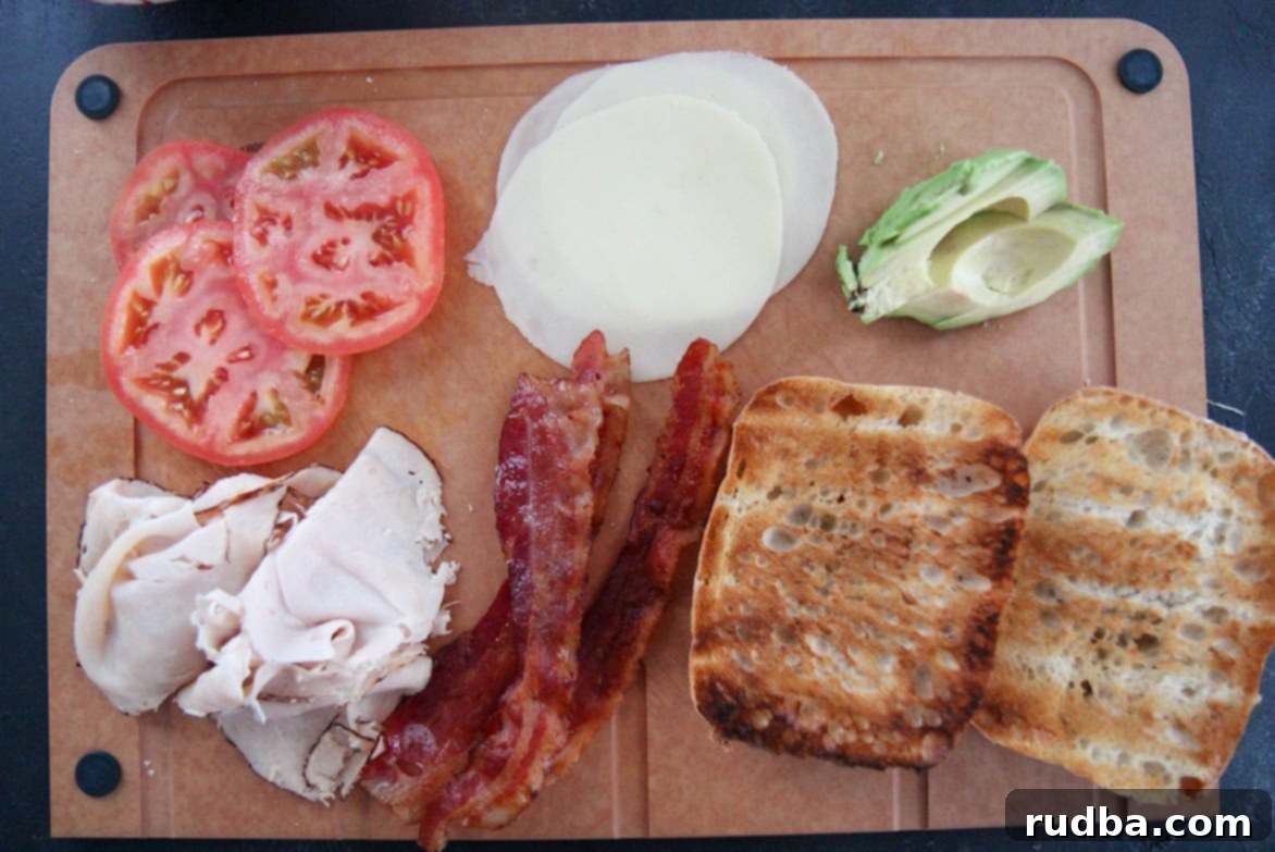 A wooden cutting board displaying all the fresh, delicious components for a turkey bacon avocado sandwich: sliced tomatoes, crispy bacon, savory turkey, provolone cheese, creamy avocado slices, and toasted ciabatta bread.