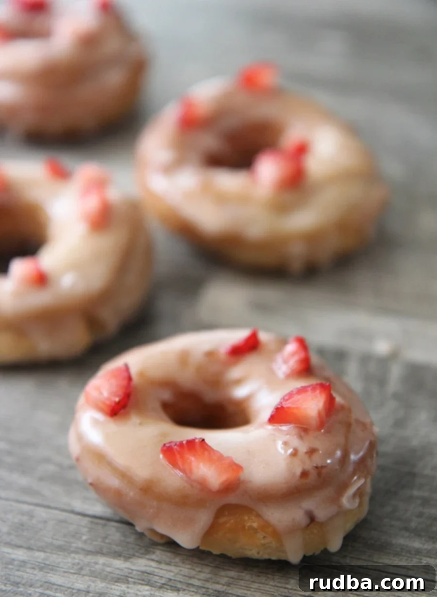 Four fresh strawberry donuts on a gray surface, garnished with strawberry pieces.