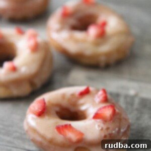 Strawberry Donut Bliss 9 Four fresh strawberry donuts on a gray surface, garnished with strawberry pieces.