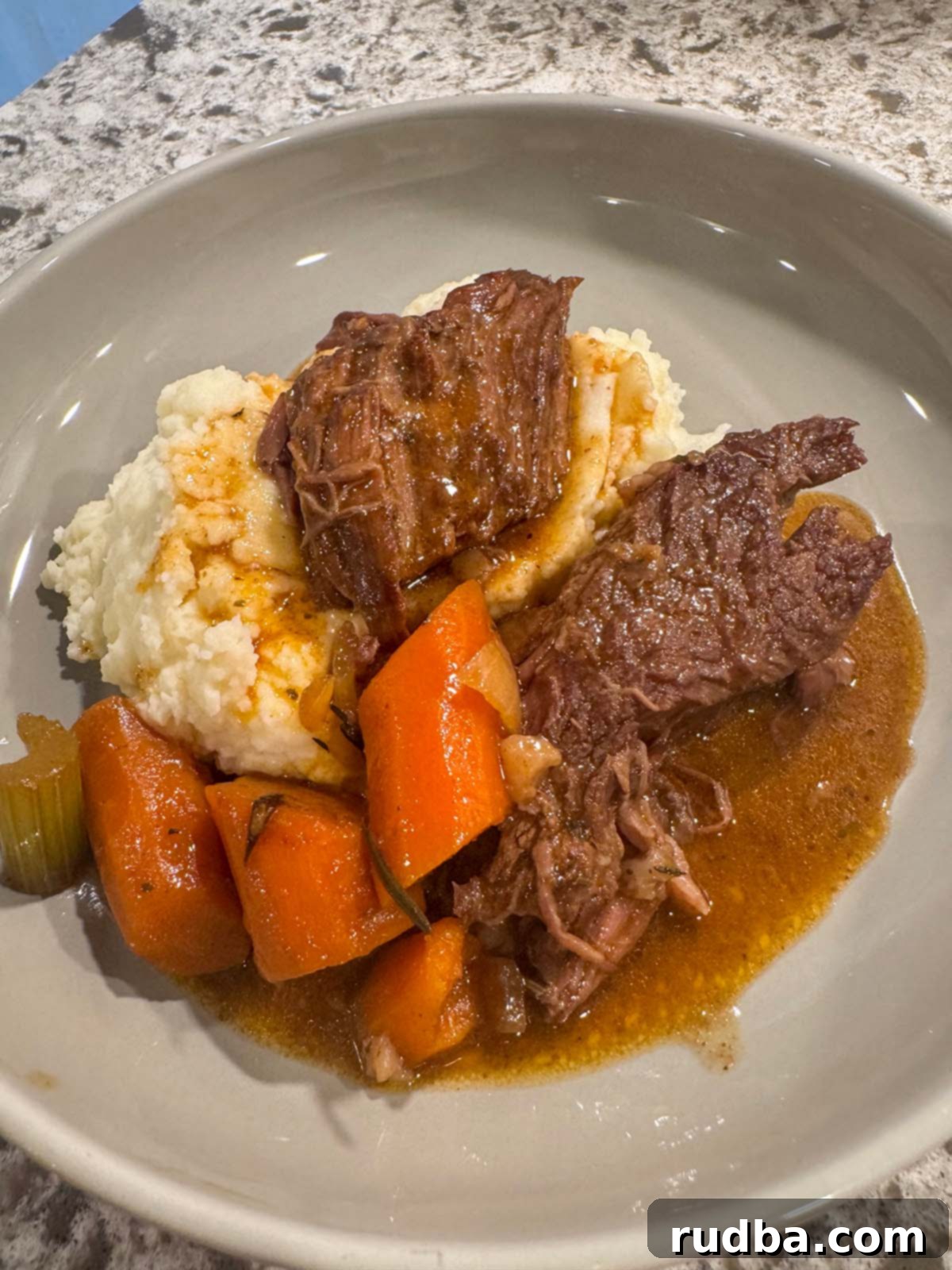 A beautifully plated slow cooker pot roast surrounded by tender carrots, celery, and rich gravy, served alongside a generous portion of creamy goat cheese mashed potatoes. This dish is the epitome of comfort food, perfect for a cozy meal.