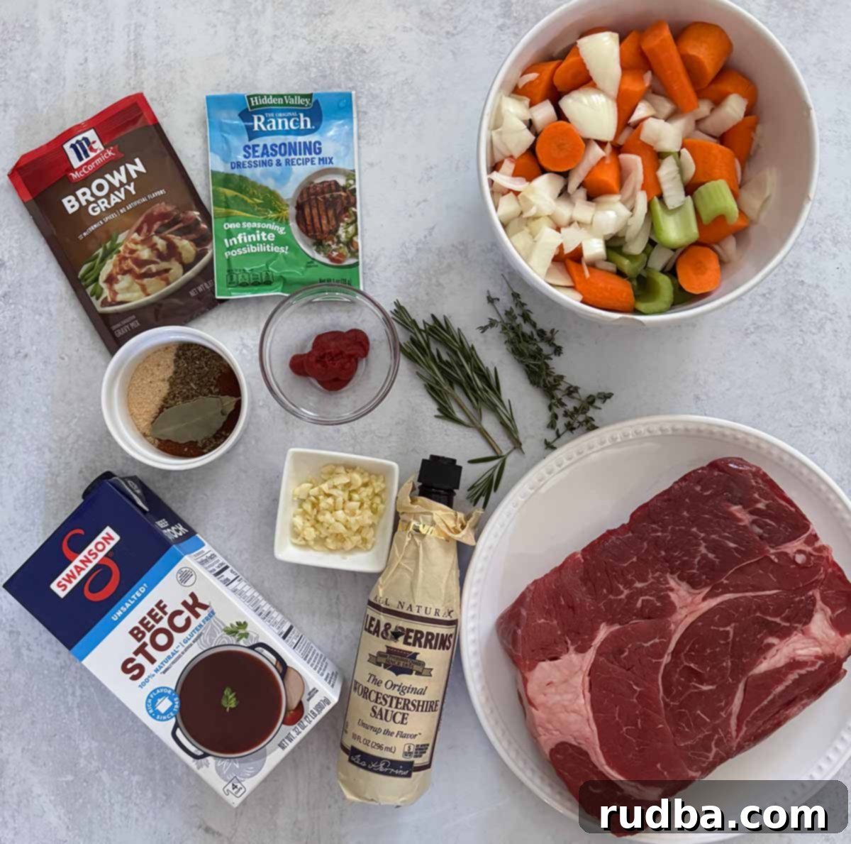 An organized assortment of fresh ingredients laid out for pot roast preparation, including a raw chuck roast, whole carrots, celery stalks, a yellow onion, garlic cloves, and various essential spices and herbs.