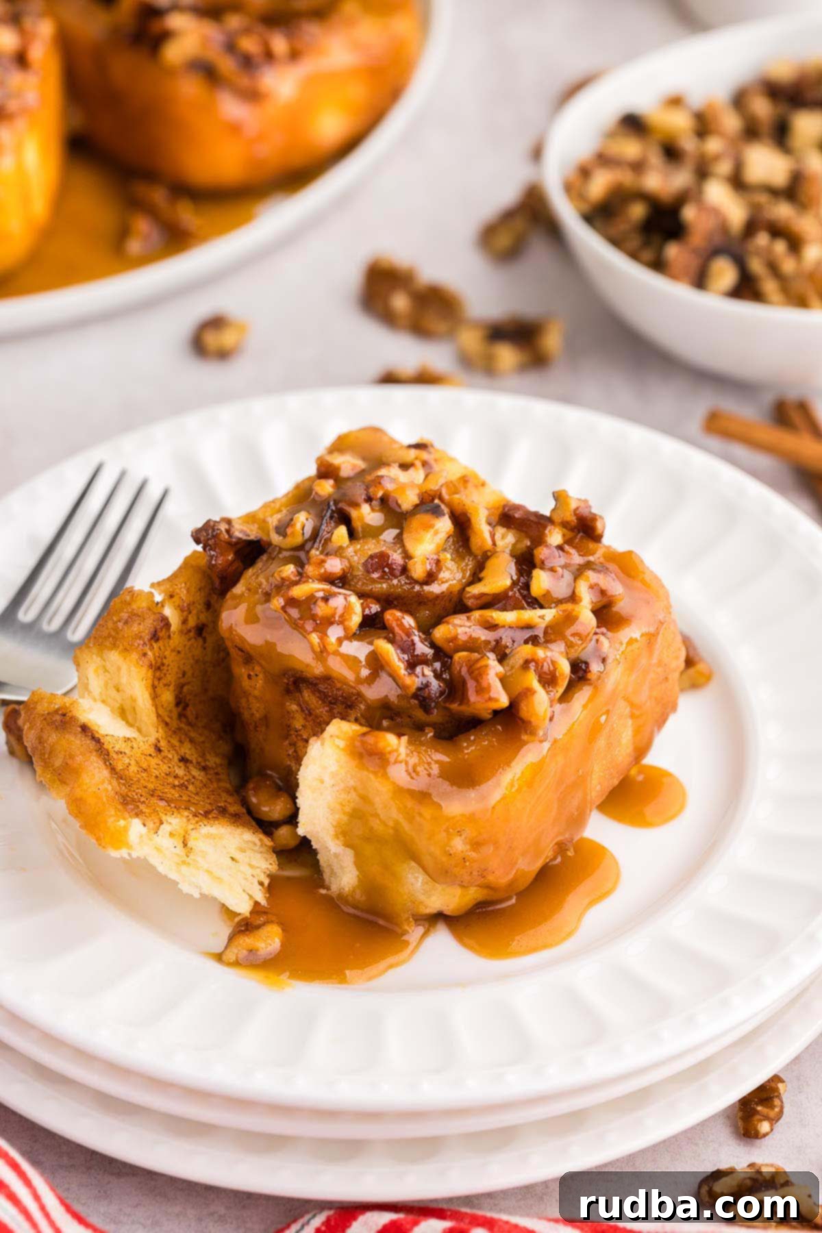 A single perfectly glazed sticky bun on a pristine white plate, accompanied by a fork, showcasing its soft interior and rich caramel topping with visible pecans.